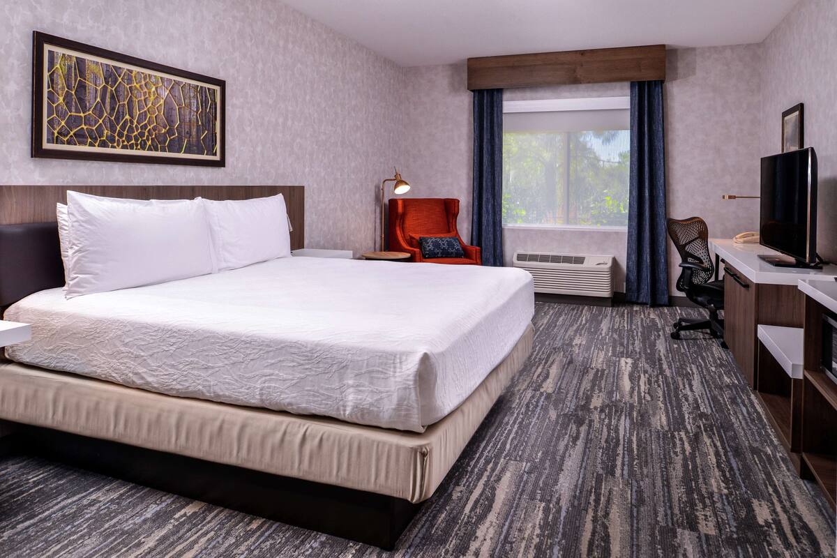 Quarto de hotel com uma cama grande, uma escrivaninha com computador, uma cadeira, uma televisão e uma janela com cortinas.