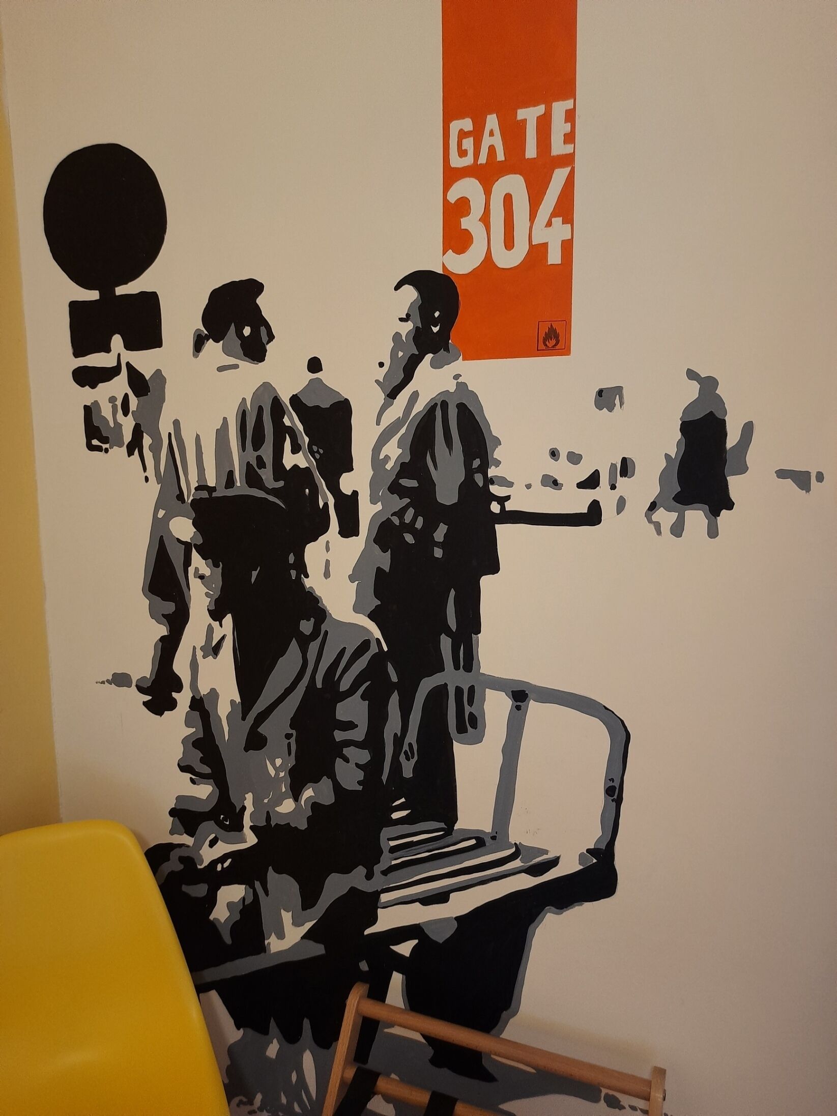A room with a white wall, a yellow chair, and a black and white mural depicting people and a chair.