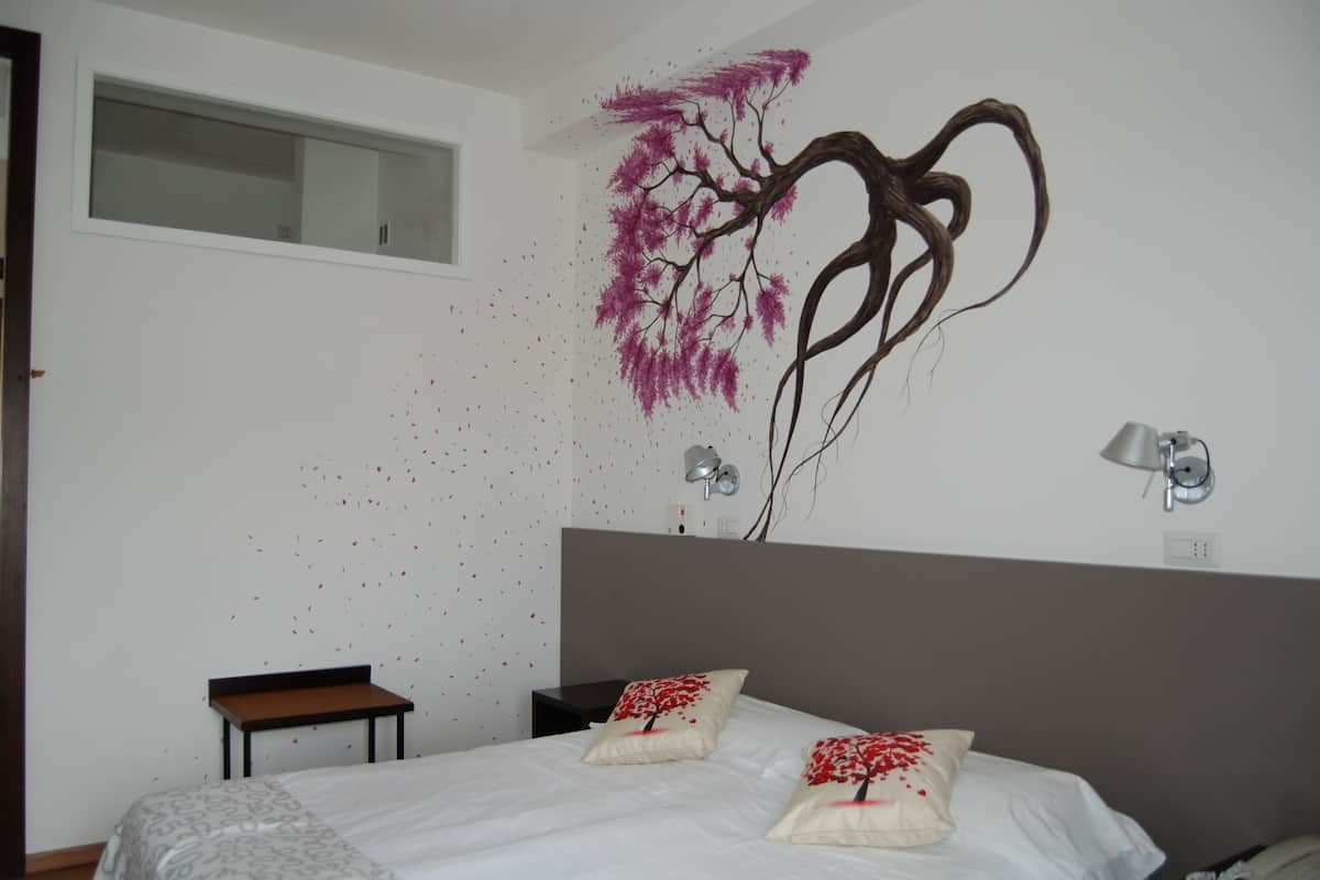 A bedroom with a large bed, a nightstand, a small table, and a wall-mounted lamp.