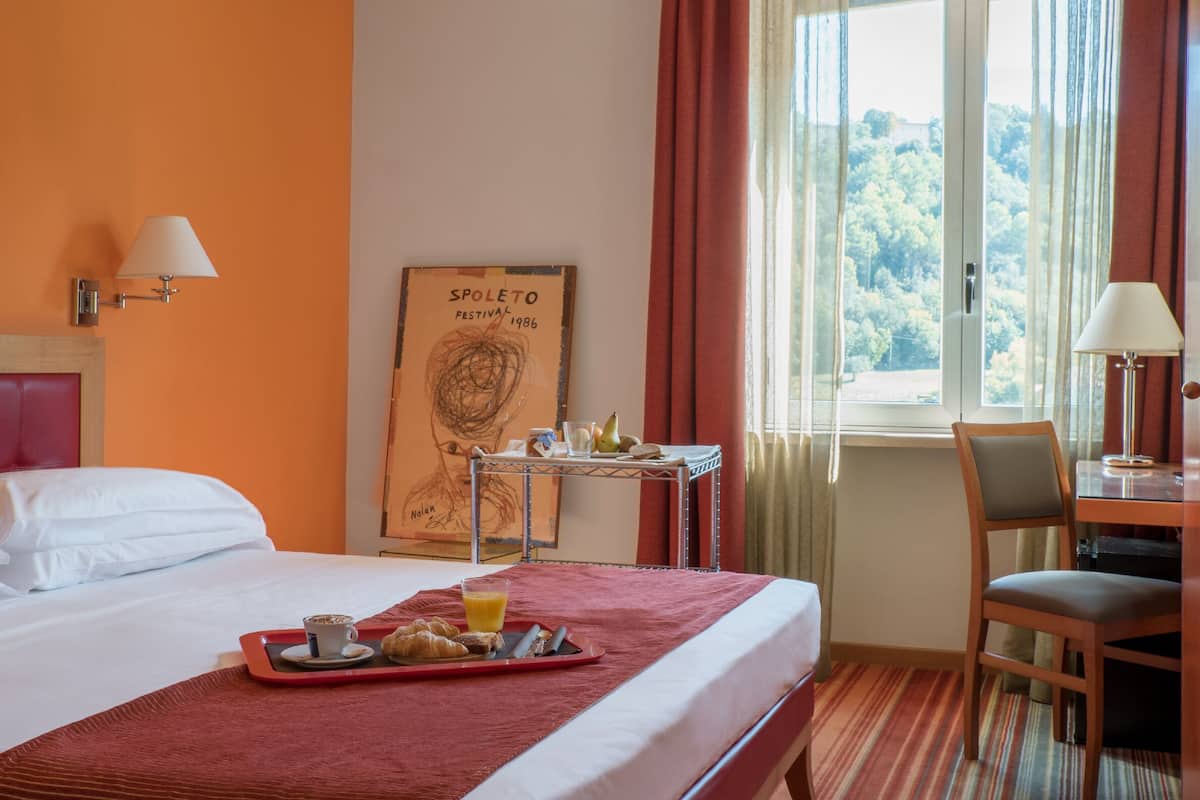 A hotel room with a bed, breakfast tray, a small table with a lamp, a chair, and a window with curtains.