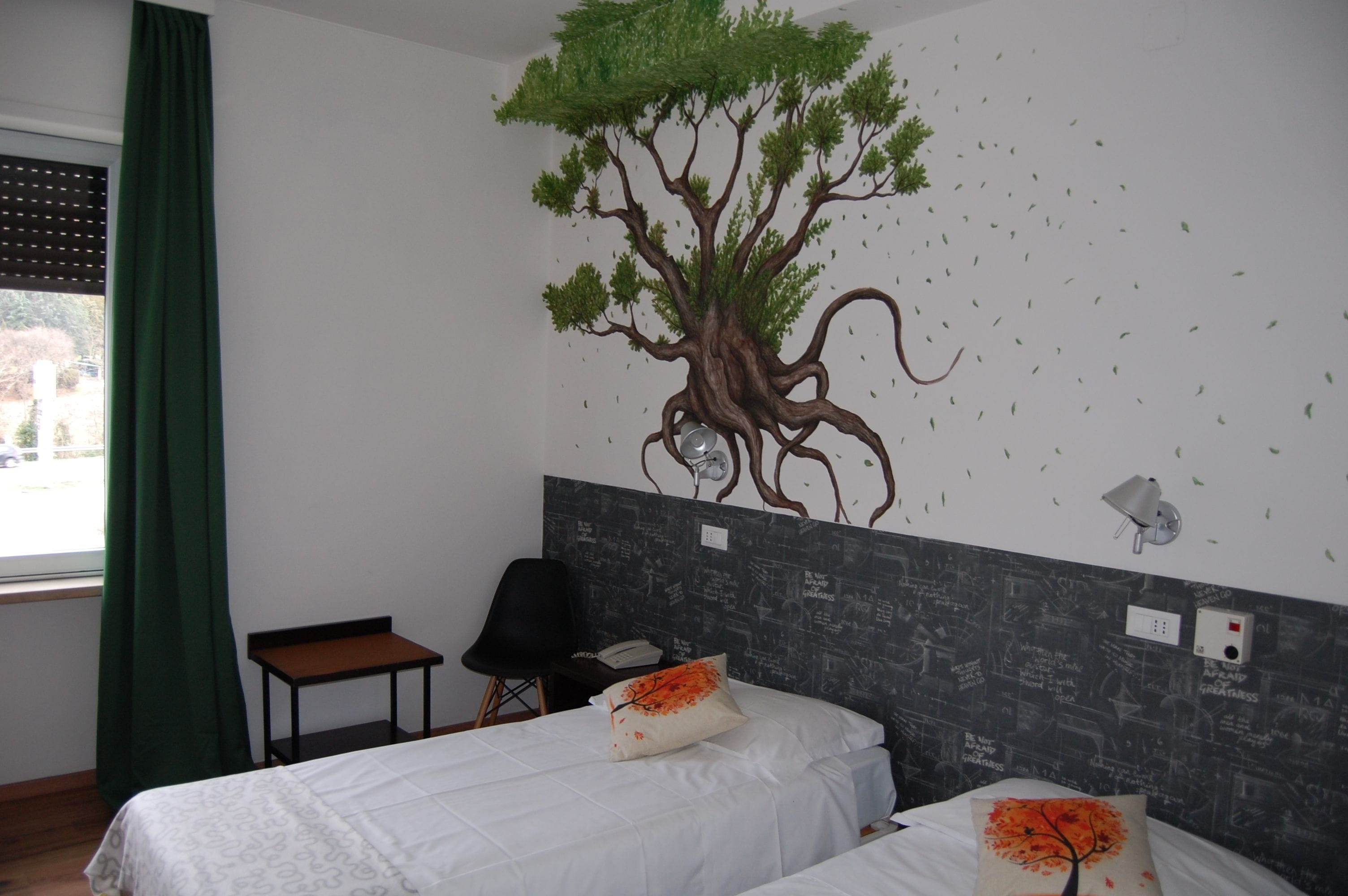 Quarto de hotel com duas camas, cortina verde, adesivo de árvore na parede, uma pequena mesa e uma cadeira.