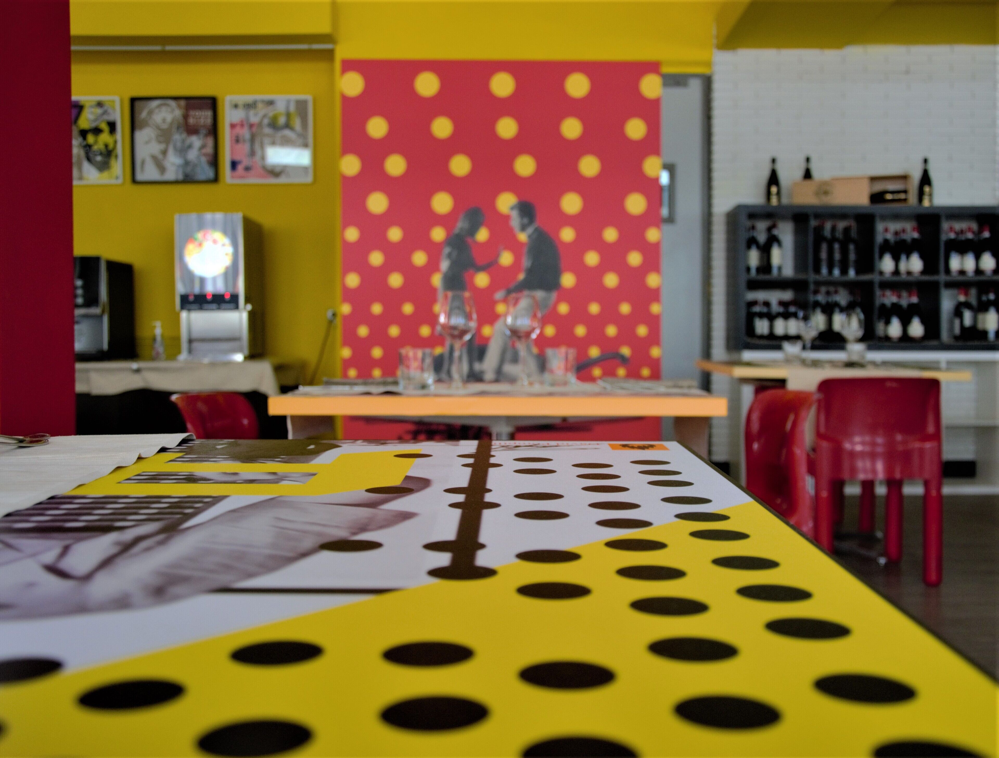 Uma área de refeições com uma mesa coberta por um tecido estampado de bolinhas, cadeiras vermelhas e uma parede com padrão de bolinhas.