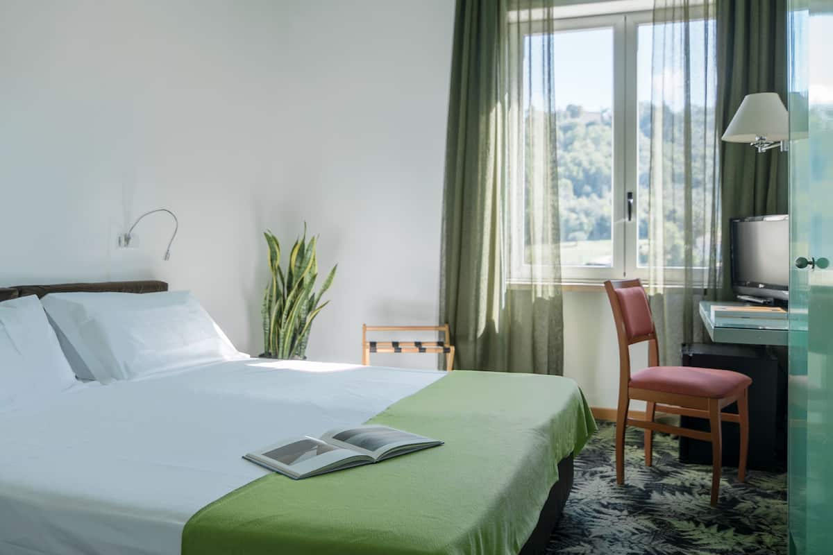 A neatly made bed with a green bedspread, a small table with a lamp, a chair, and a potted plant in a hotel room with a view of greenery outside.