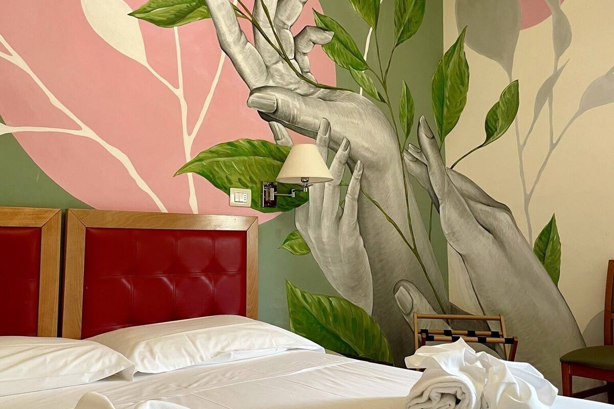 A hotel room with a large wall mural of a tree, a red headboard, a bed with white linens, and a small bedside table with a lamp.