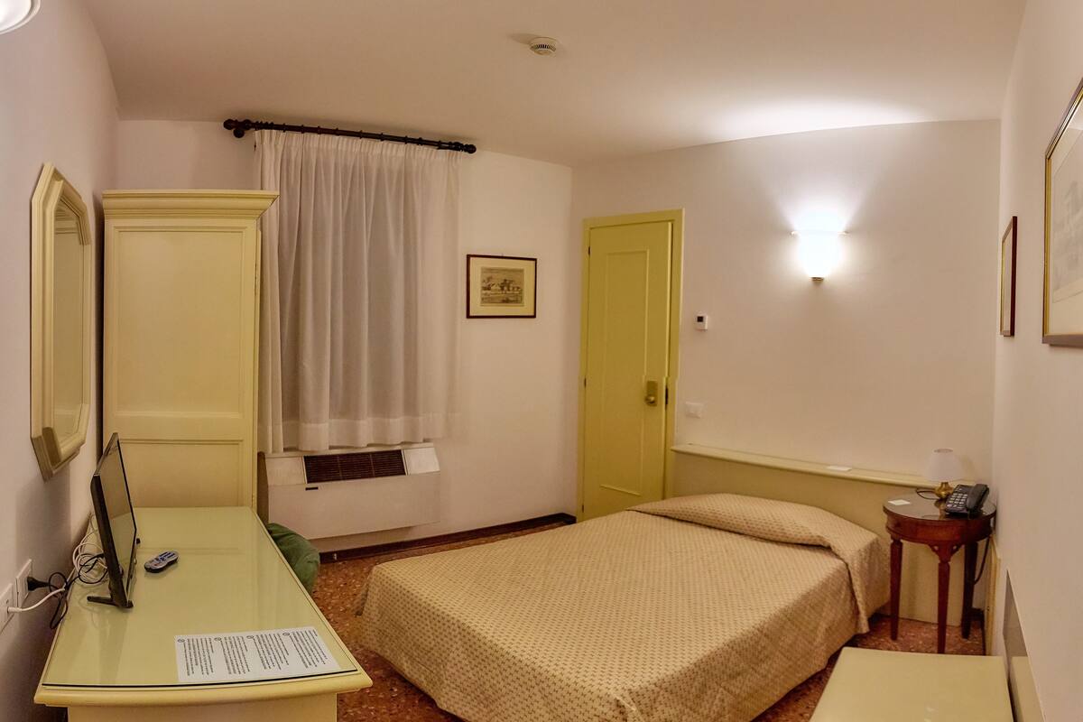 A small, simple room with a bed, a desk, a chair, a bedside table, a mirror, a window with curtains, a door, and a wall-mounted light.