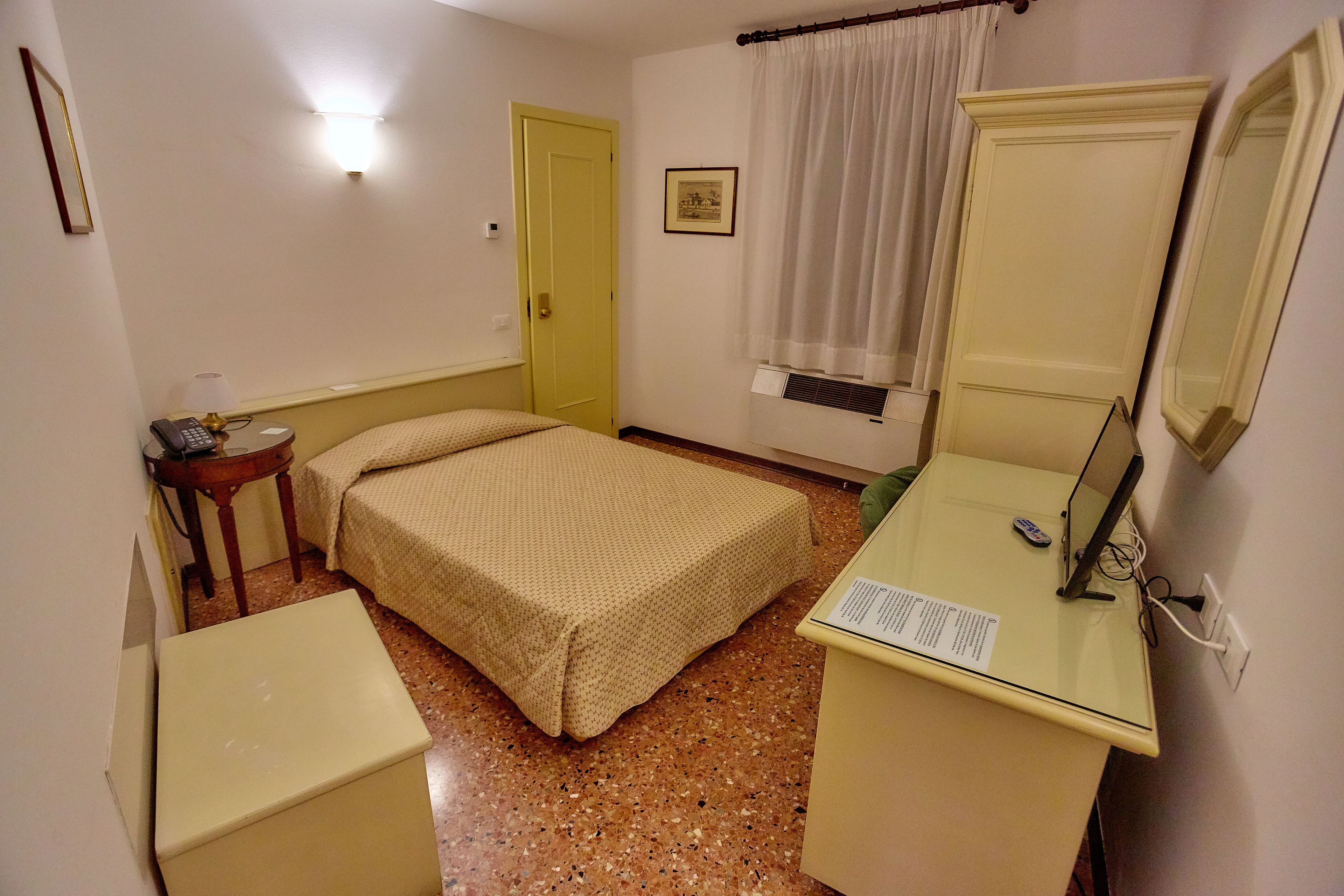 A small, compact room with a bed, a desk with a mirror, a bedside table, and a door.