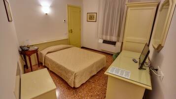 A small, compact room with a bed, a desk with a mirror, a bedside table, and a door.
