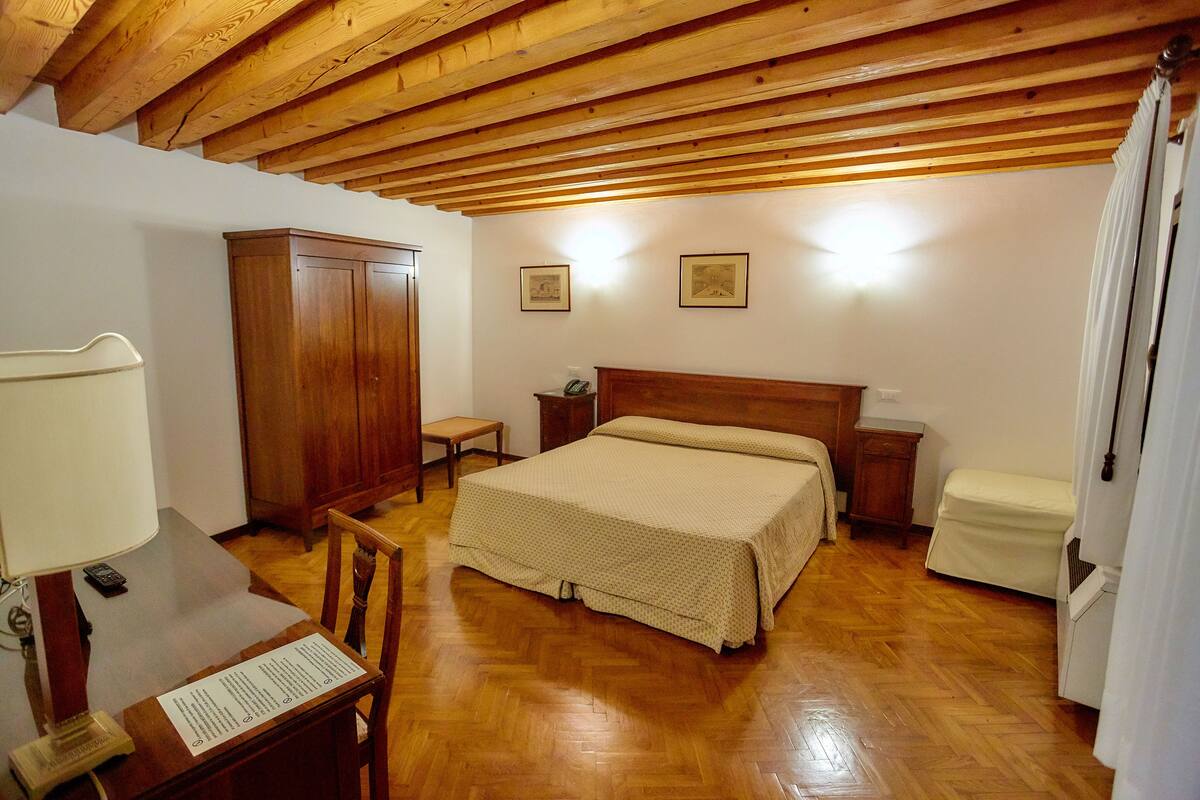 A bedroom with a wooden ceiling, a bed, a nightstand, a bedside table, a chair, a wardrobe, and a desk with a lamp.