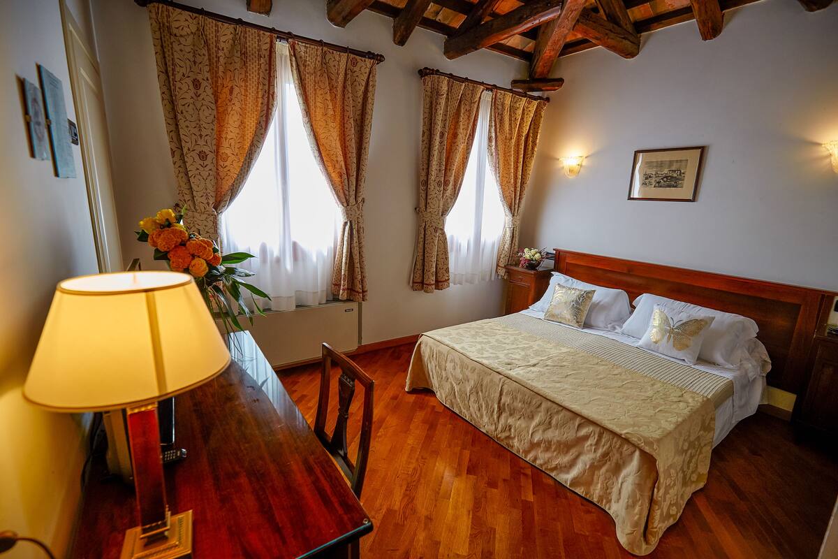 A bedroom with a wooden bed, a desk with a lamp, and two windows with curtains.