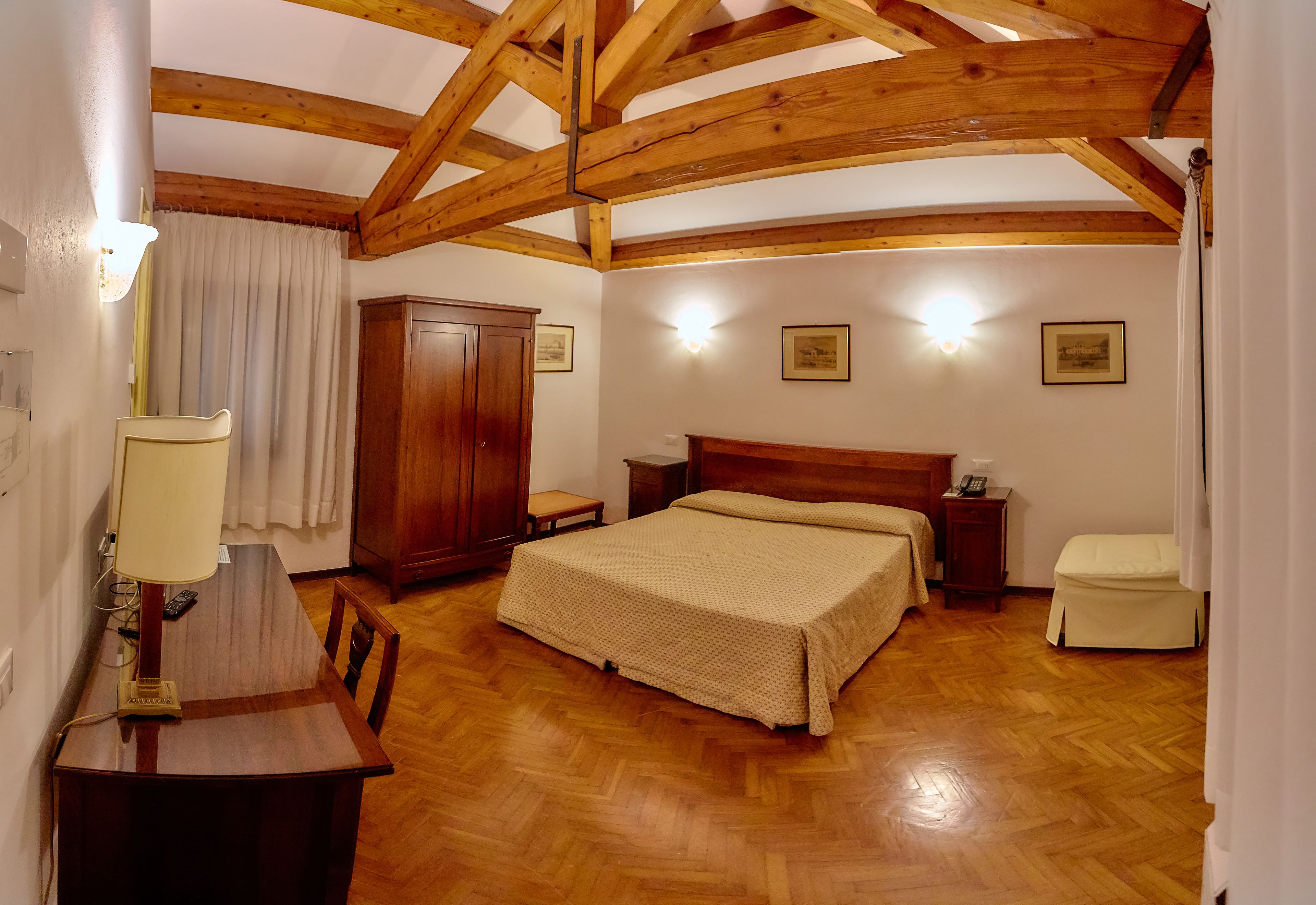 A bedroom with a wooden ceiling, a bed, a bedside table, a nightstand, a wardrobe, and a desk with a lamp.