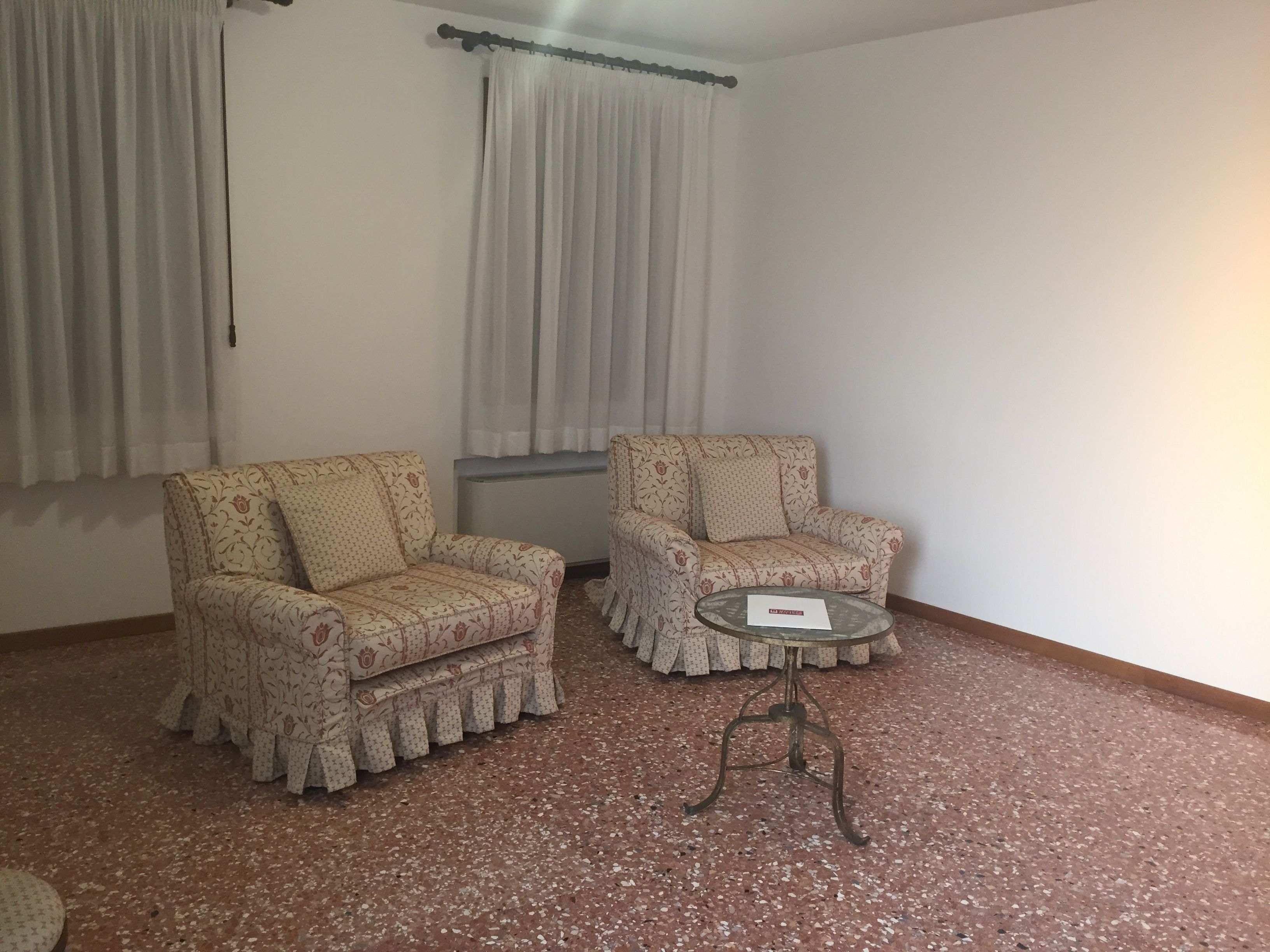 A room with two patterned armchairs and a small round table, a window with curtains, and a tiled floor.