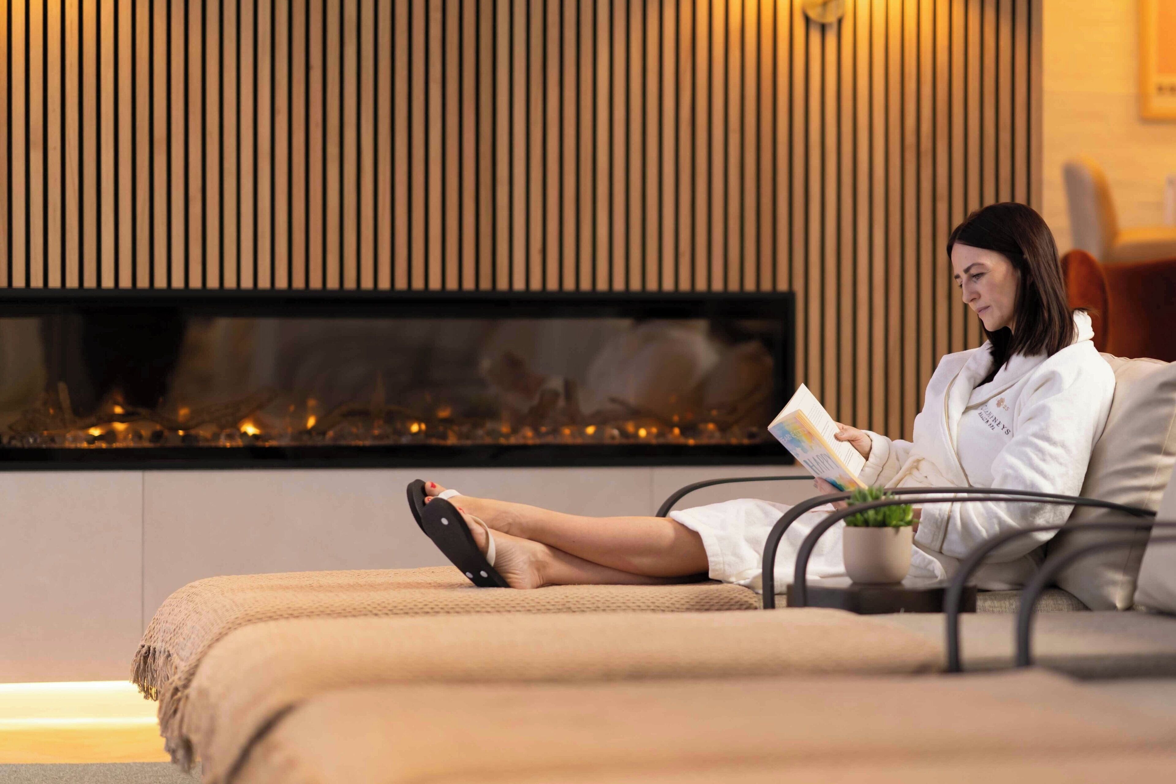 Una persona sentada en un sofá leyendo un libro en una habitación con una chimenea moderna y una planta en maceta.
