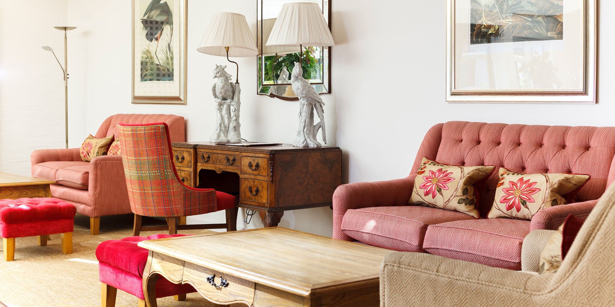 A living room with a red sofa, a plaid armchair, a wooden coffee table, and a matching ottoman. There is a wooden side table with a lamp and a decorative sculpture.