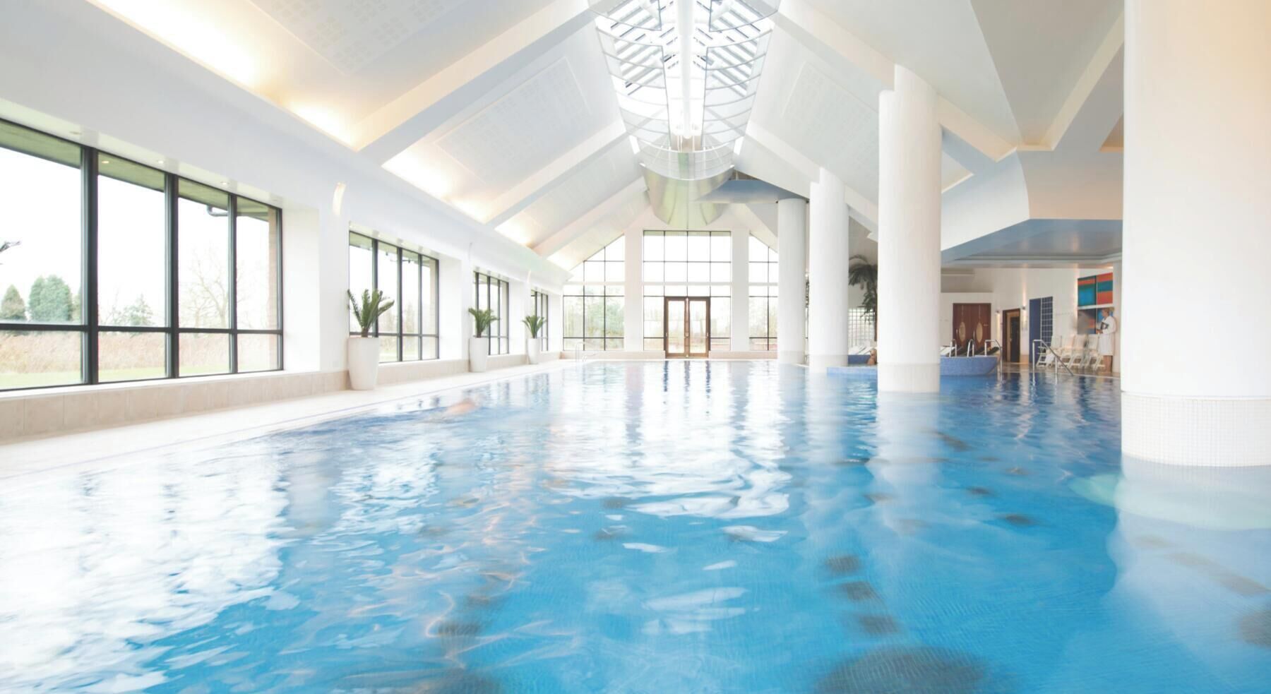 Une piscine intérieure avec des parois en verre transparent, de grandes fenêtres et un plafond haut doté de lucarnes.