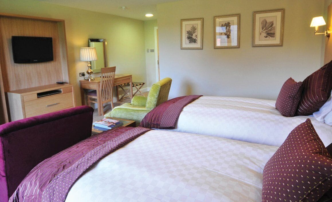 A hotel room with two beds, a desk, a chair, a TV, and framed artwork on the wall.