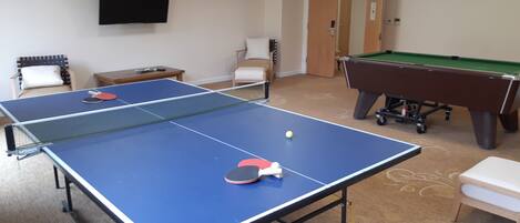Una habitación con dos mesas de ping pong, una mesa de billar y un televisor en la pared.