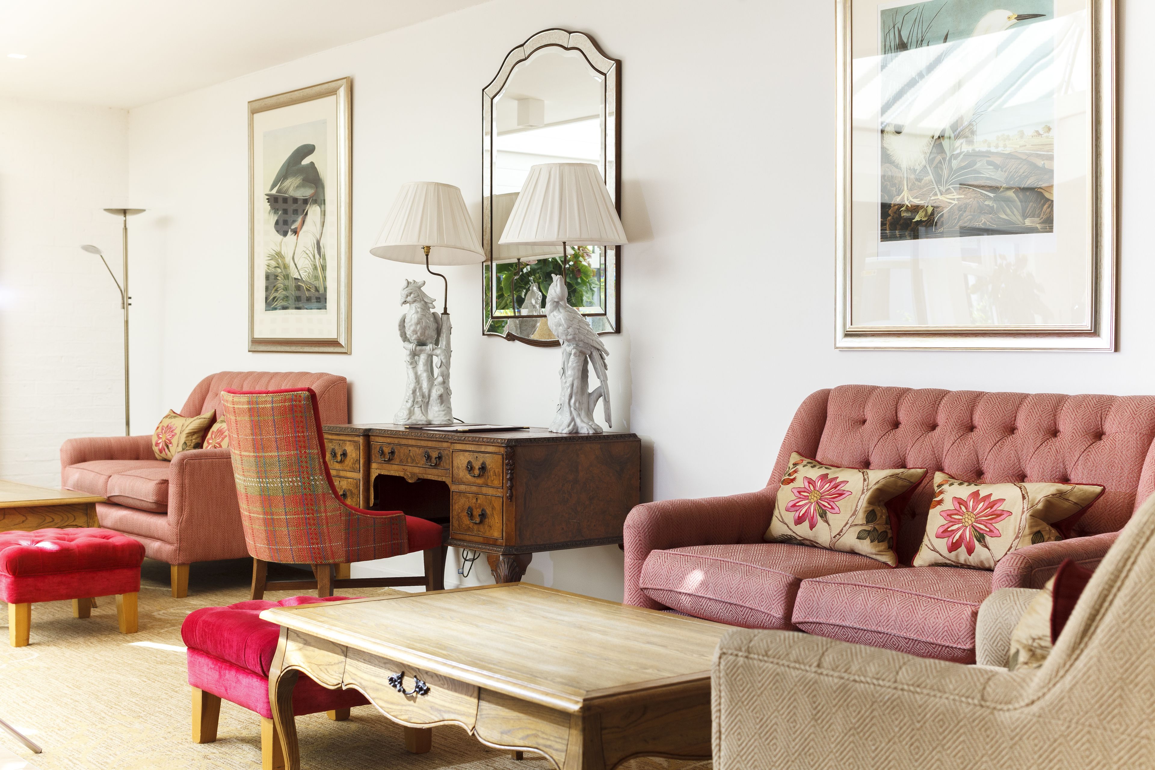 A living room with a pink sofa, a red armchair, a wooden coffee table, and a matching ottoman. There is a wooden cabinet with drawers and a mirror above it.