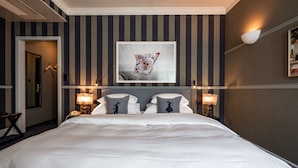 Une chambre d’hôtel avec un grand lit, un papier peint à rayures, une œuvre d’art encadrée au-dessus du lit, des lampes de chevet et une porte donnant sur une autre pièce.