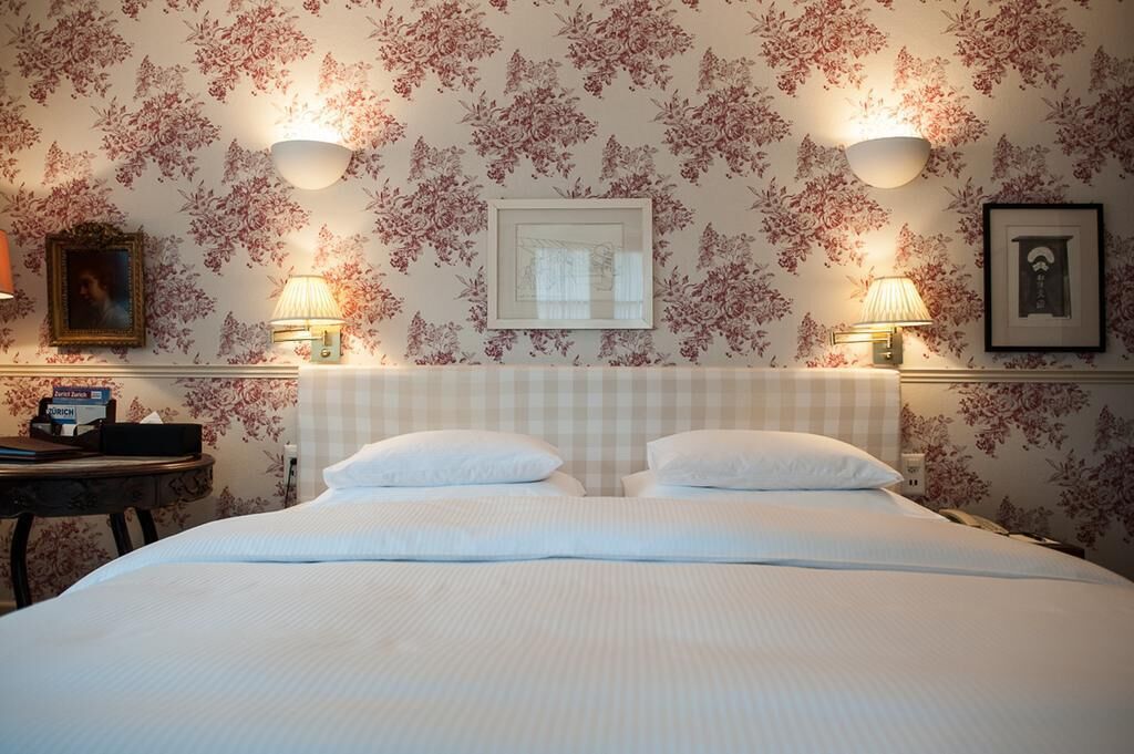 A neatly made bed with a checkered headboard, two bedside lamps, and floral wallpaper.