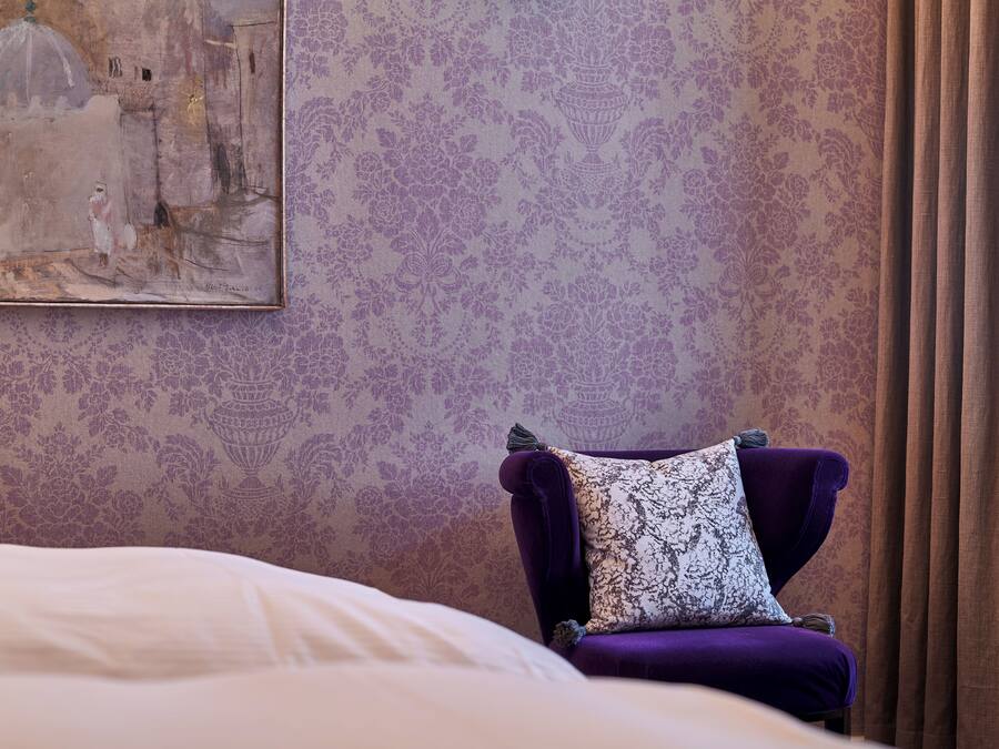 Una stanza con un letto, una poltrona viola con un cuscino decorato e una carta da parati floreale.