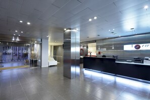 Een moderne hotelontvangst met een strakke receptiebalie, een grote pilaar en een glazen wand met de naam van het hotel.