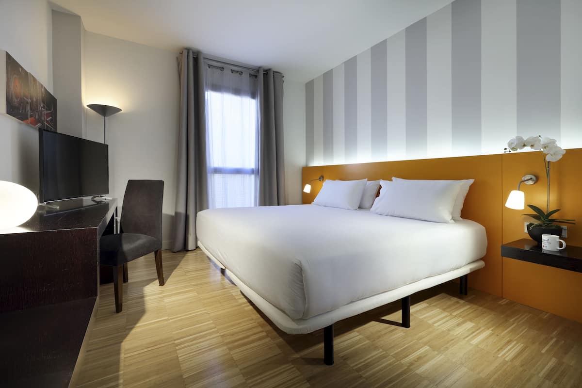 Ett hotellrum med en säng, ett skrivbord, en stol, en tv, en lampa och en krukväxt.