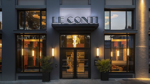 L’entrée d’un hôtel, avec une façade moderne, de grandes fenêtres en verre et une enseigne bien visible indiquant « LE CONTI ».