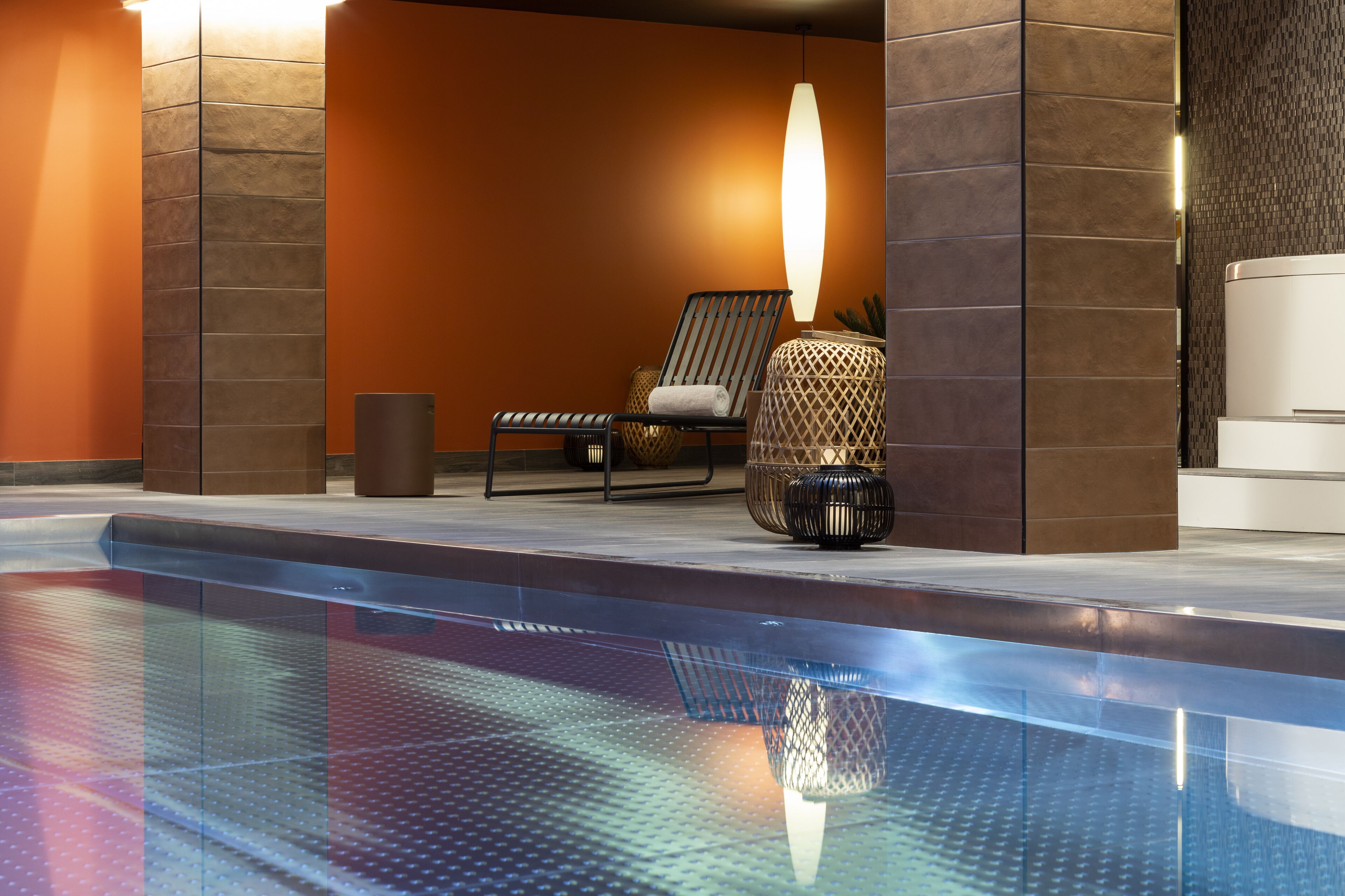 A modern indoor pool area with a lounge chair, a decorative lamp, and a reception desk.