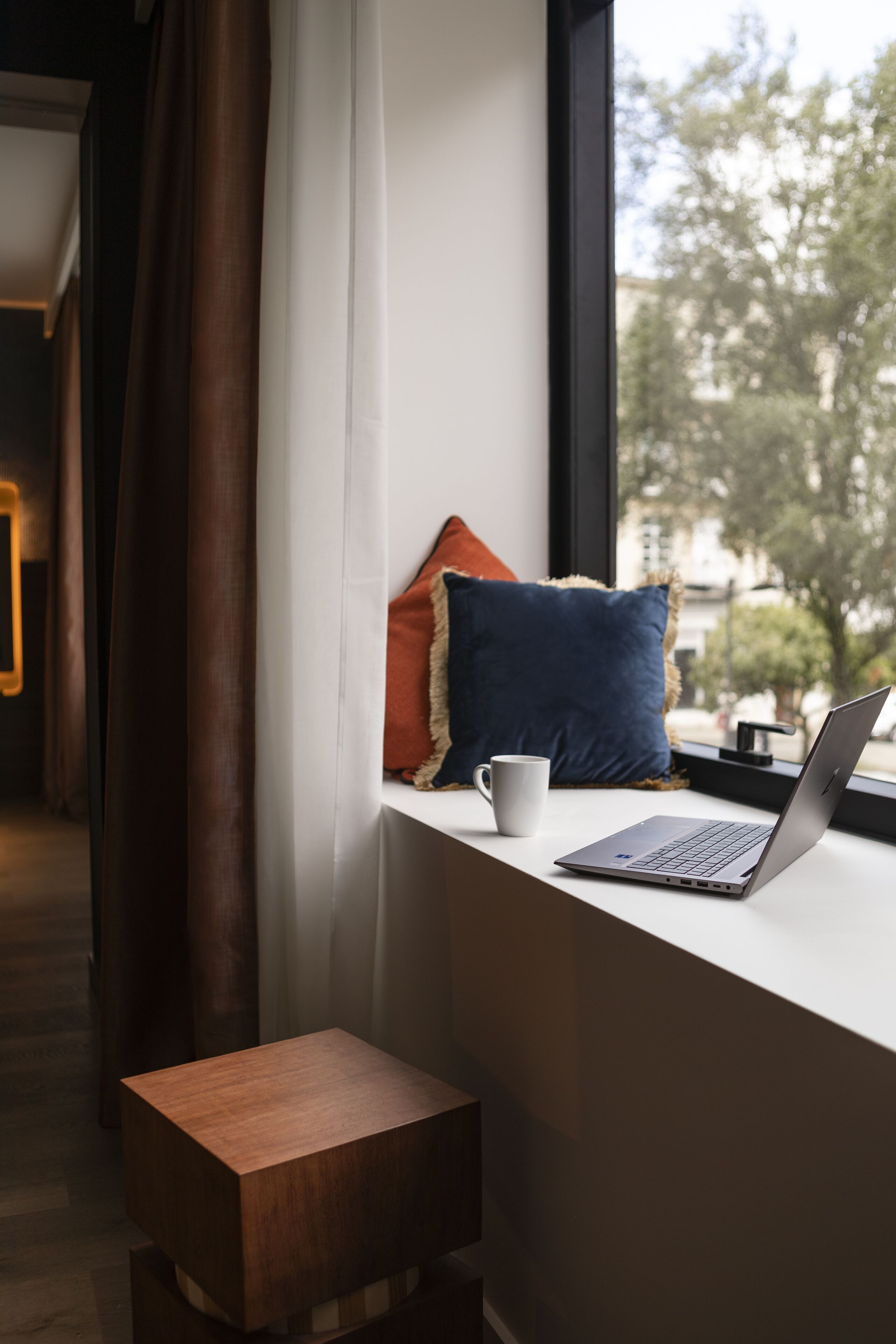 A laptop on a window sill with a white mug beside it, a wooden stool, and a view of trees and buildings outside.