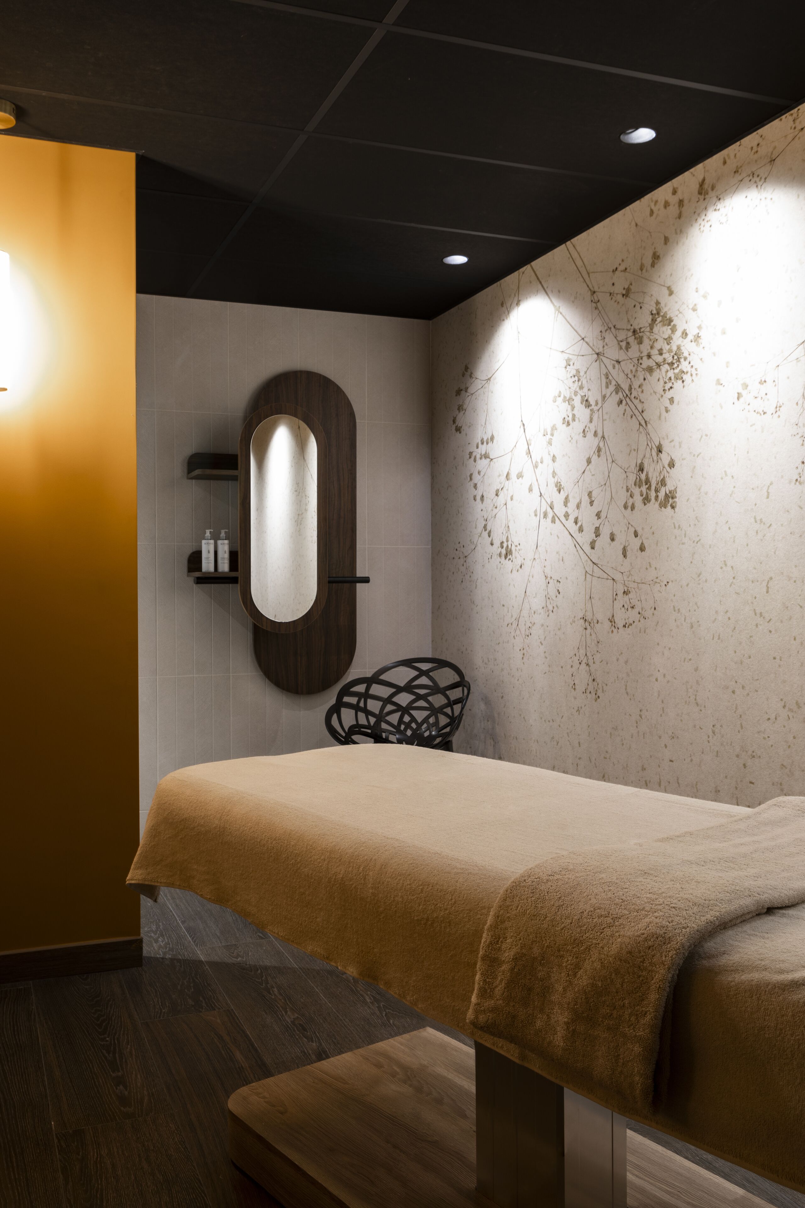 Een moderne spa-ruimte met een houten bankje, een wandspiegel en een decoratieve wand met een bladmotief.