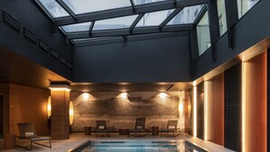 Une piscine intérieure moderne dotée d’un toit en verre, de chaises longues et d’un mur en pierre.