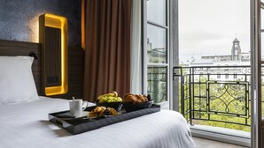 Une chambre d’hôtel avec un lit, un plateau contenant des articles pour le petit-déjeuner, une fenêtre donnant sur des immeubles et un miroir décoratif.