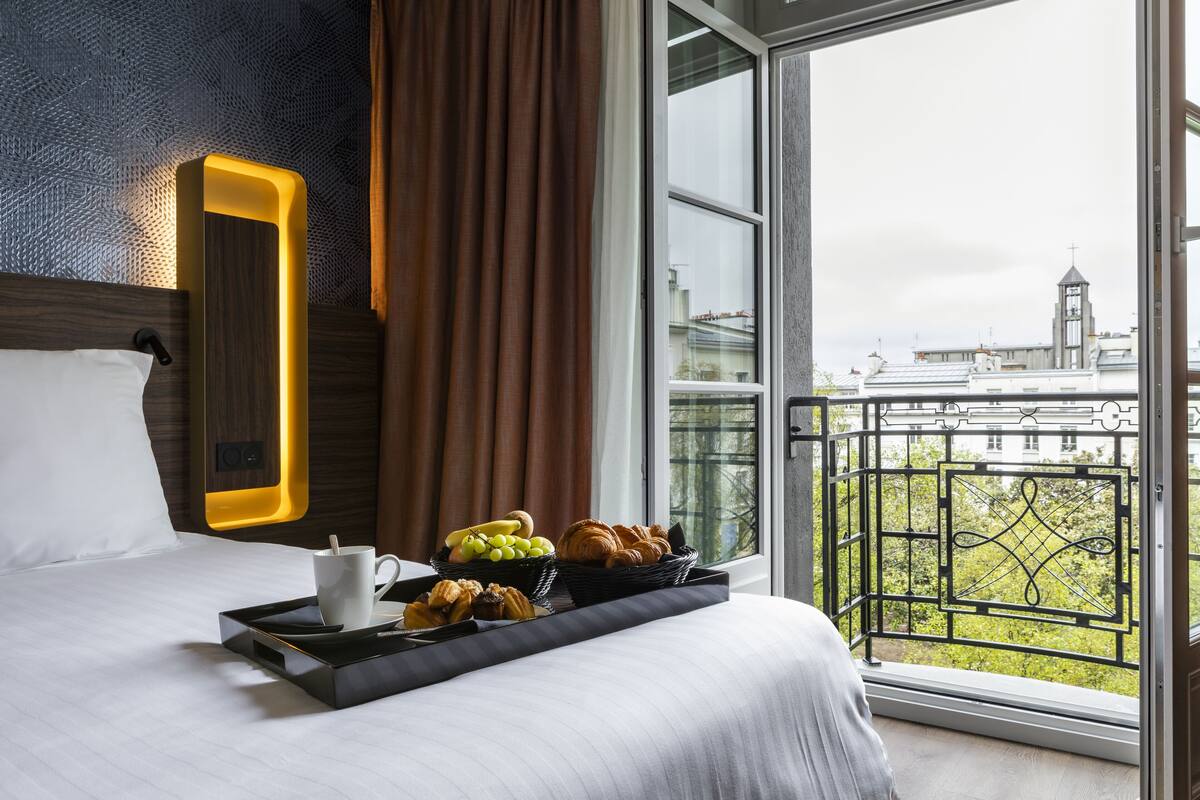 Una camera d'albergo con un letto, un vassoio con prodotti per la colazione, una finestra vista edifici e uno specchio decorativo.