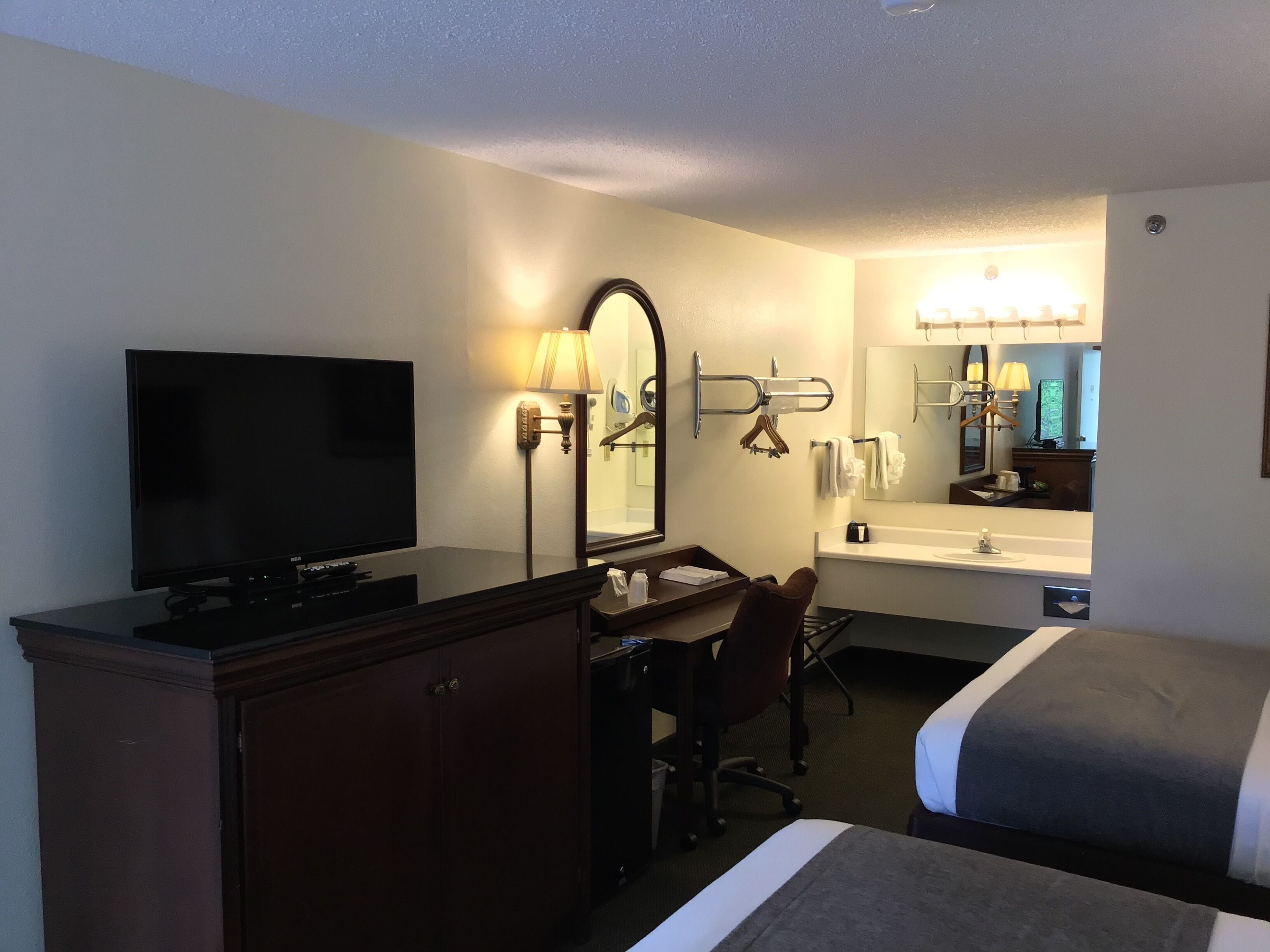 Habitación de hotel con televisor de pantalla plana, escritorio con silla, cama, cómoda, espejo y baño con lavamanos y toallero.
