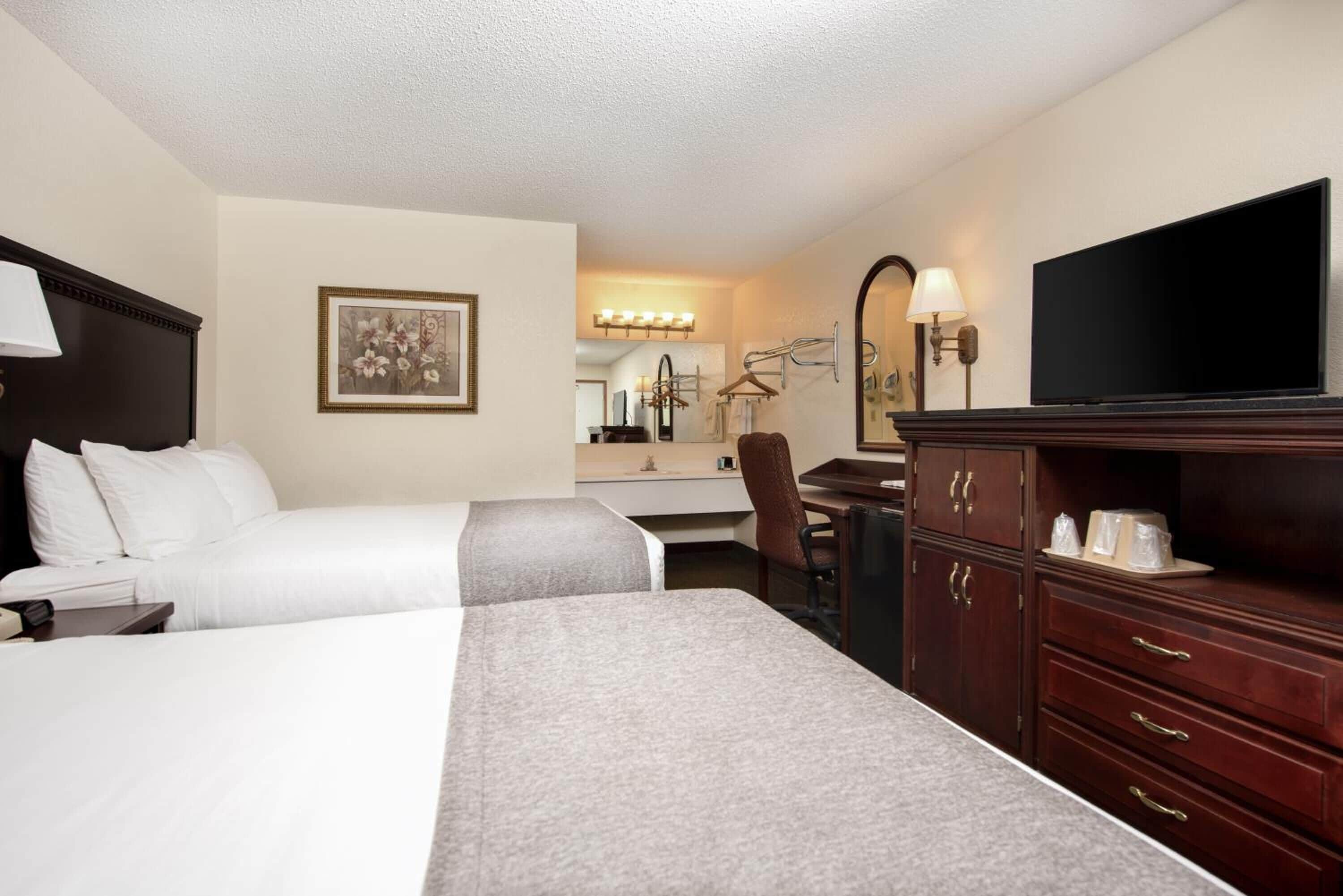 Une chambre d’hôtel avec deux lits, un bureau, une chaise, une commode, une télévision et une salle de bain équipée d’un miroir et d’un éclairage.