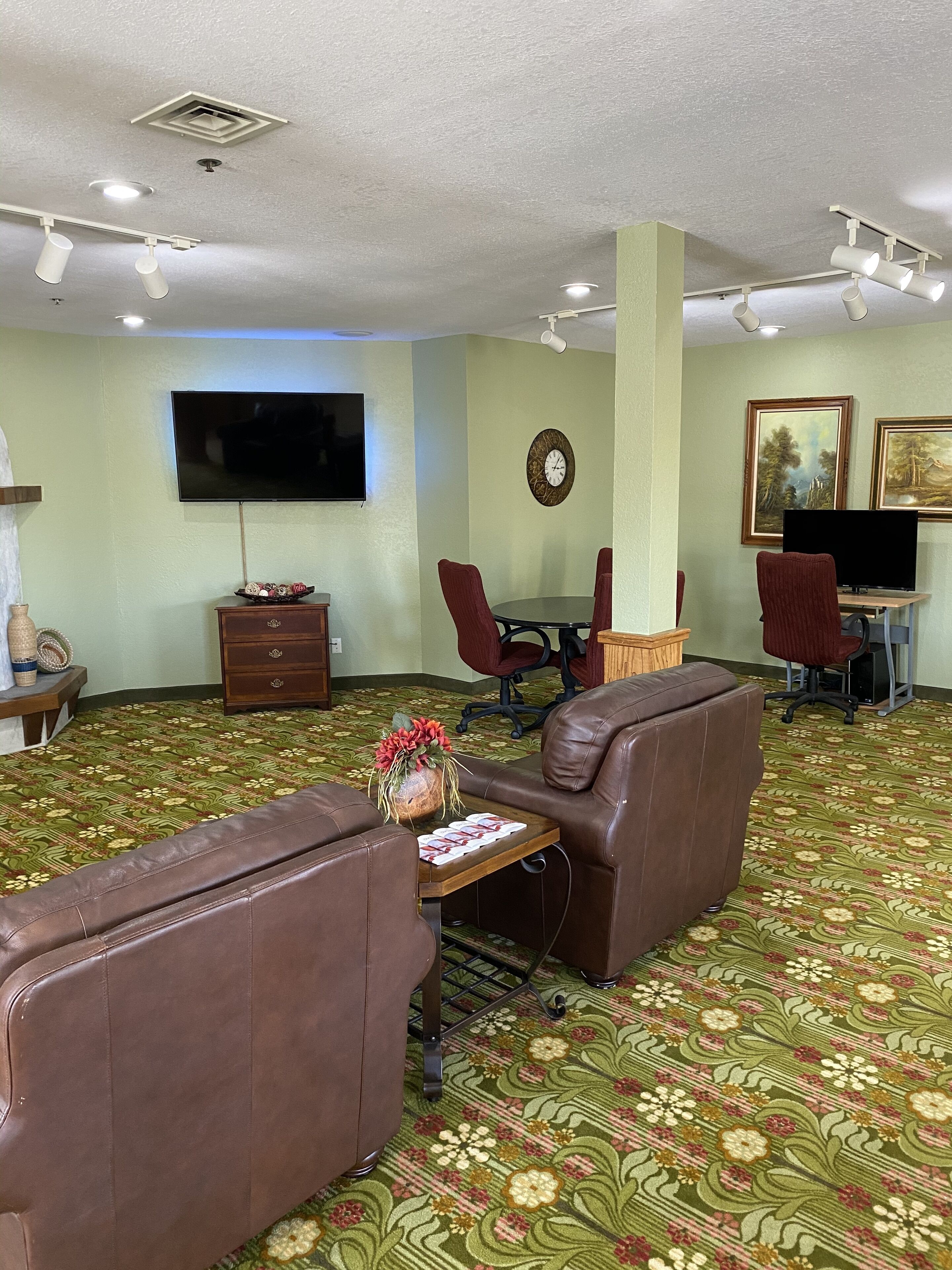 A living room with a patterned carpet, leather sofas, a wooden coffee table, a TV mounted on the wall, and a dining area with chairs and a table.