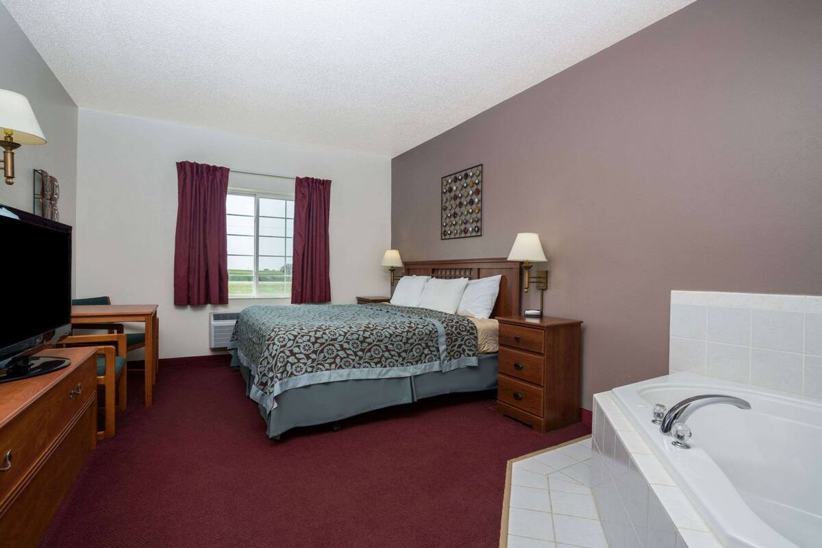 A hotel room with a bed, a desk with a TV, a bedside table with lamps, a bathroom with a bathtub, and a window with curtains.