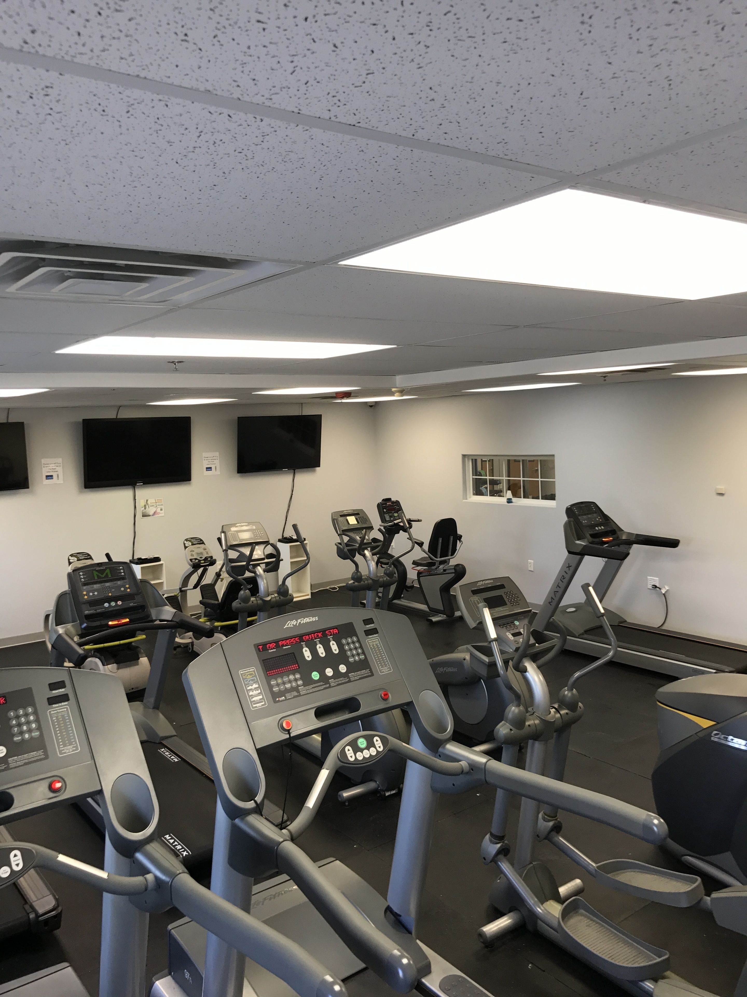 A gym with various exercise machines, including treadmills and elliptical trainers, and a large TV screen on the wall.