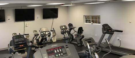 A gym with various exercise machines, including treadmills and elliptical trainers, and a large TV screen on the wall.