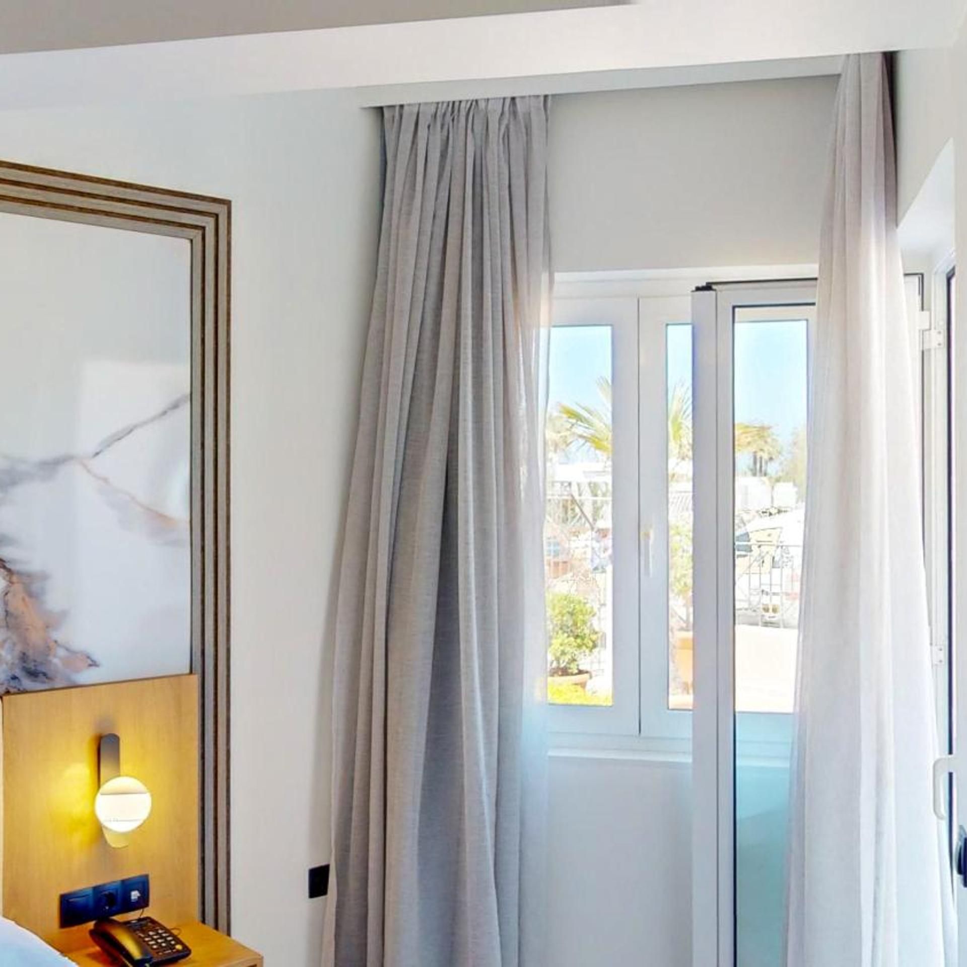A hotel room with a bed, a bedside table with a lamp, a window with curtains, and a view of the outside.