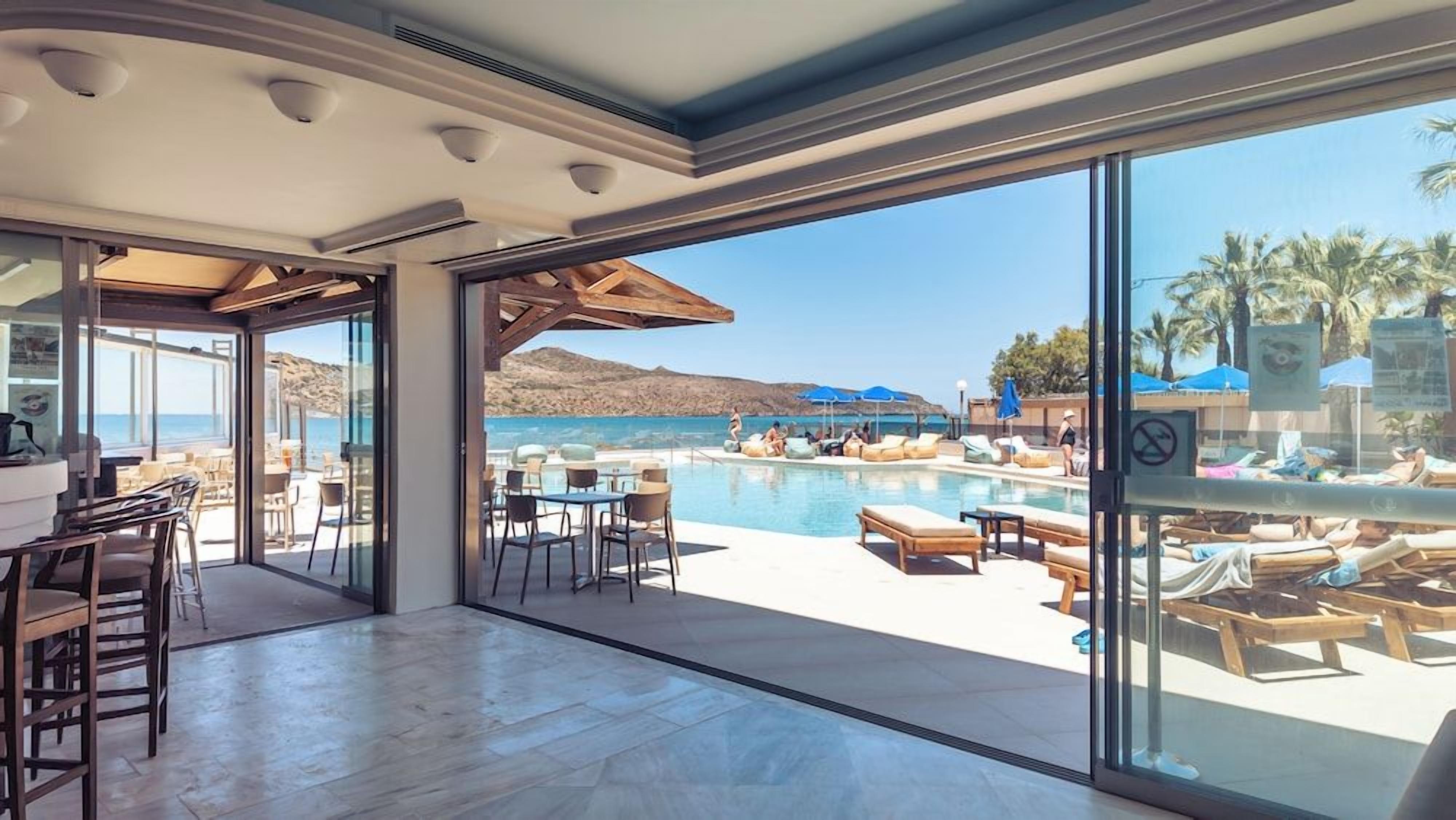 Une chambre d’hôtel moderne dotée d’une grande baie vitrée donnant sur un espace piscine avec des chaises longues et des parasols.