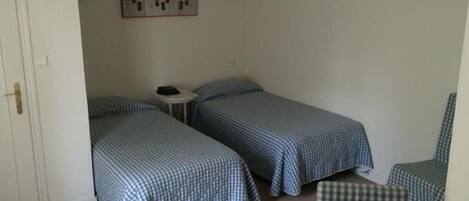 A small, simple room with two single beds, a small table, and a chair.