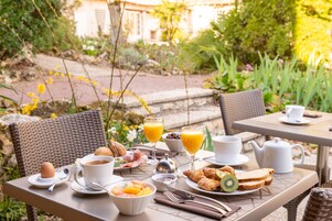 Café da manhã ao ar livre com cadeiras de vime, uma mesa com itens de café e vista para o jardim.