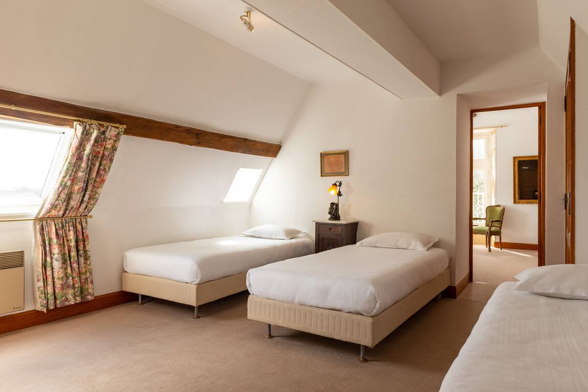 A hotel room with two beds, a window with floral curtains, a small table with a lamp, and a painting on the wall.