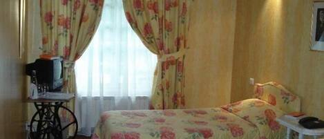 A hotel room with two beds, a small desk with a television, and a window with floral curtains.