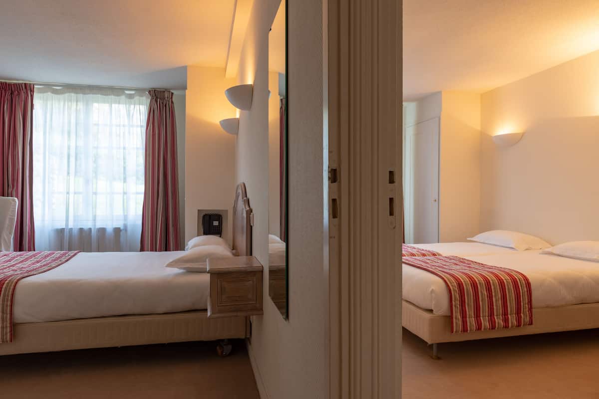 A hotel room with two beds, a bedside table, a mirror, and a window with curtains.