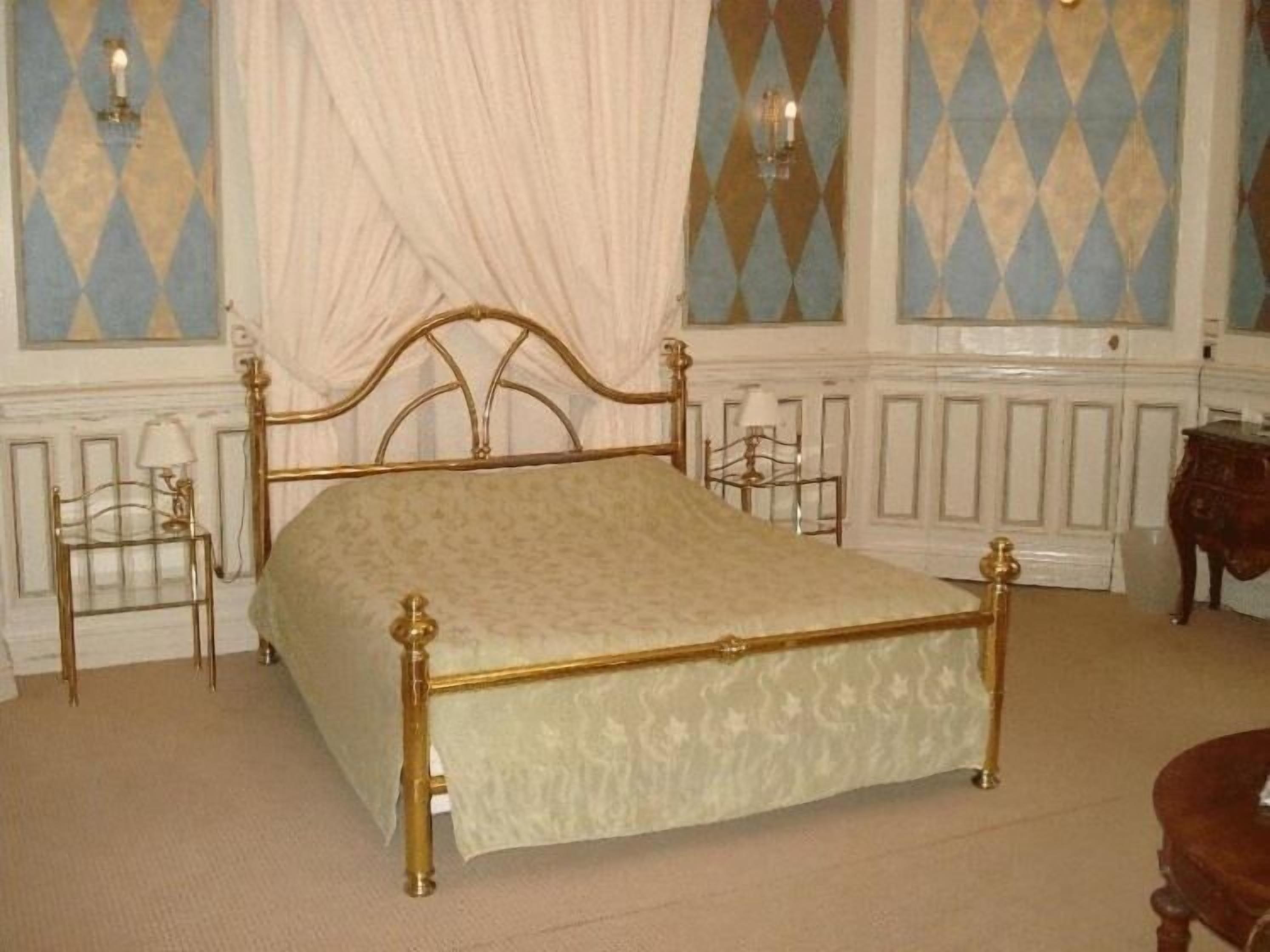 Una cama clásica con dosel y estructura dorada, con un edredón estampado, flanqueada por dos mesitas de noche con lámparas, en una habitación con papel tapiz estampado y un radiador blanco.