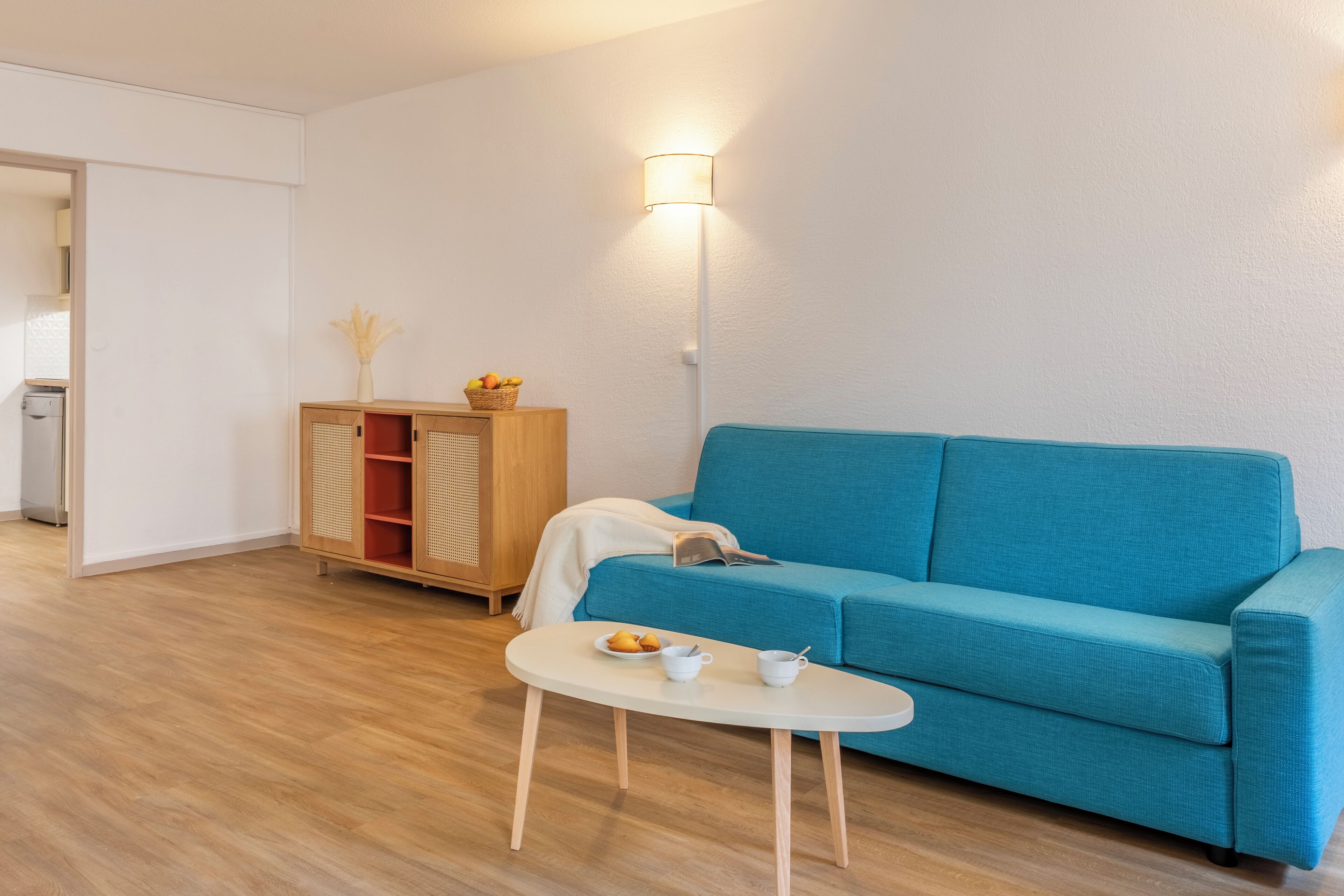 A modern living room with a blue sofa, a small white table with a cup and saucer, a wooden cabinet with a basket of fruit, and a floor lamp.