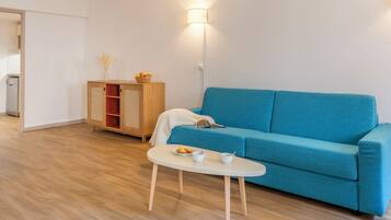A modern living room with a blue sofa, a small white table with a cup and saucer, a wooden cabinet with a basket of fruit, and a floor lamp.