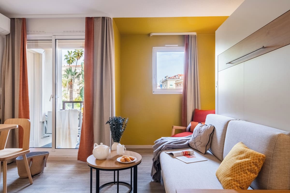 Een moderne hotelkamer met een bank, een tafeltje met een theepot en gebak, een raam met uitzicht op palmbomen, en een schuifdeur naar een terras.